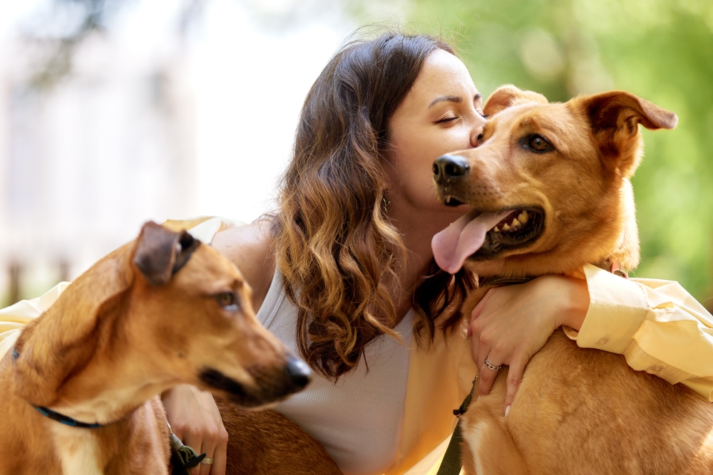 A woman embraces her two dogs with love and a gentle kiss in a sunny outdoor setting, showing deep companionship and joy.